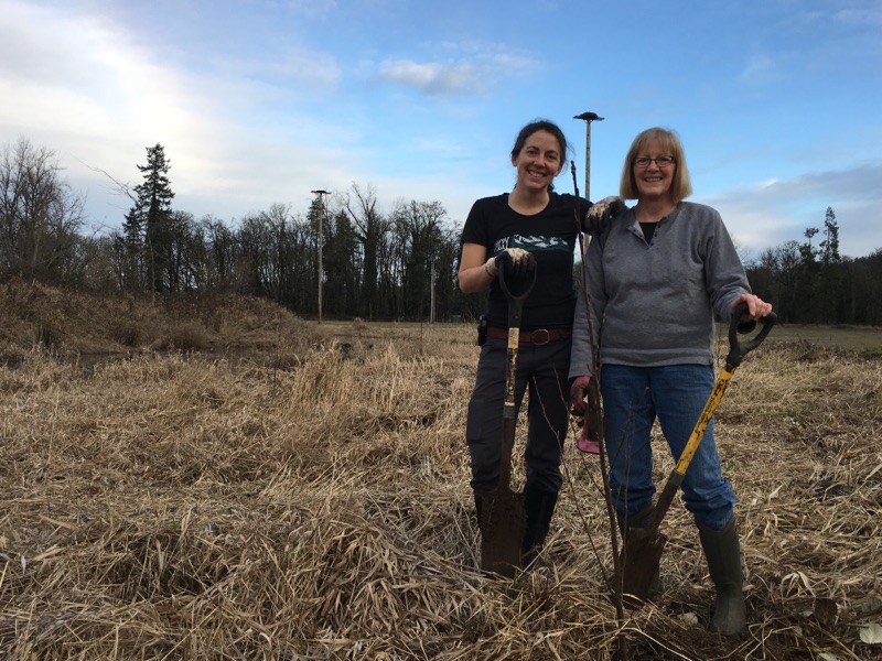 Two white women pose behind a young tree holding shovels and smiling.